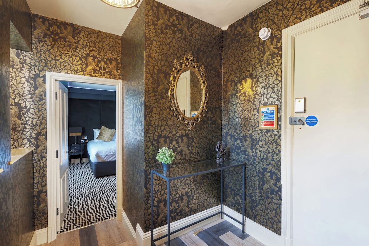 Un couloir avec un papier peint à motifs, un miroir, une porte avec une pancarte et un lit dans la pièce adjacente.