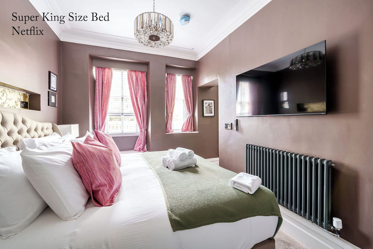 Un grand lit avec une tête de lit capitonnée, un plaid vert et des coussins roses et blancs. Un téléviseur à écran plat est fixé au mur, et un radiateur se trouve en dessous.