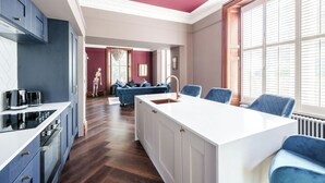 A modern kitchen with a central island, a dining area with blue chairs, and a view of a cityscape through large windows. - The Governance Apartments (Windermere)