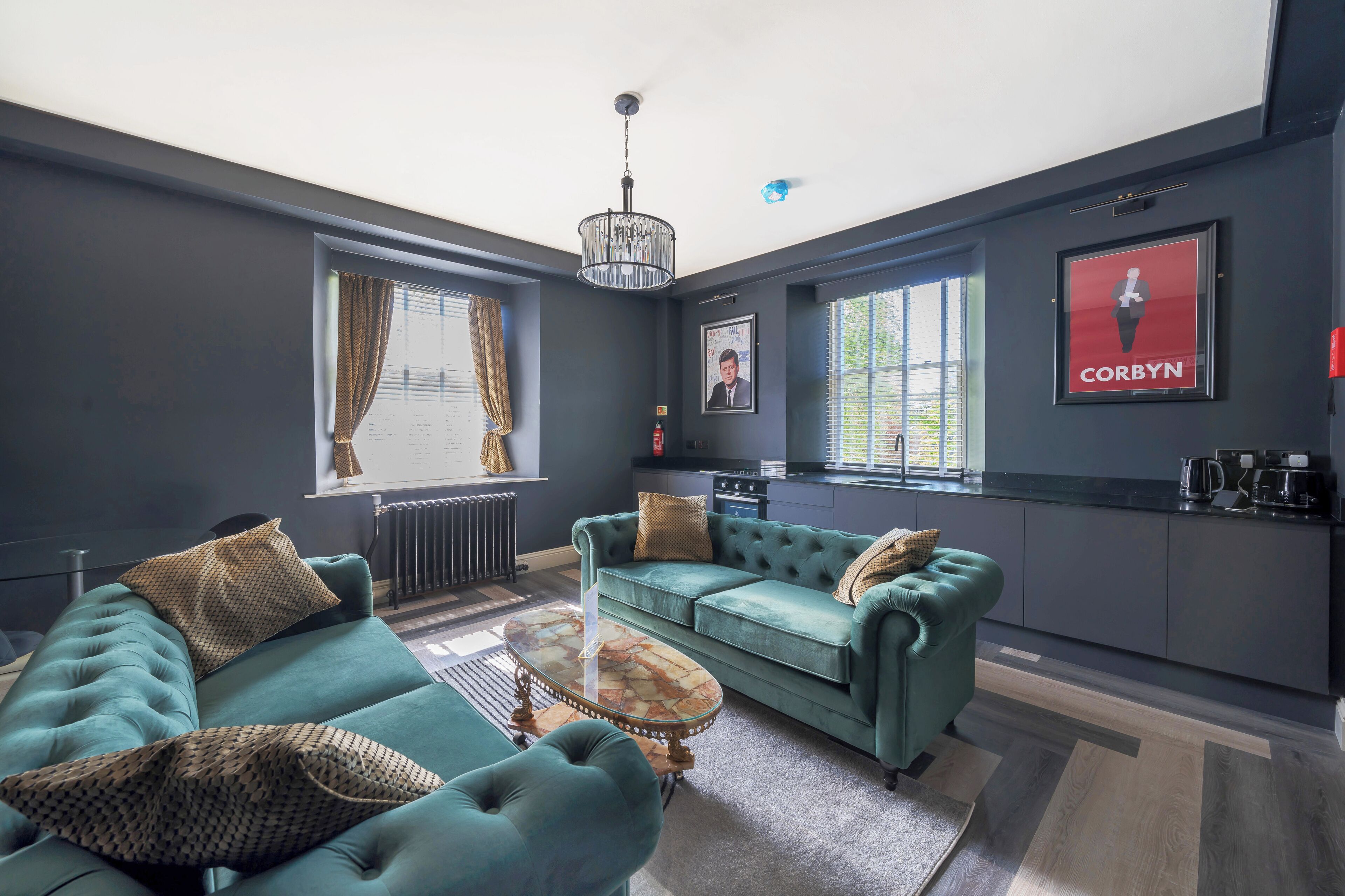 A modern living room with a teal sofa set, a coffee table, and a kitchen area visible in the background.
