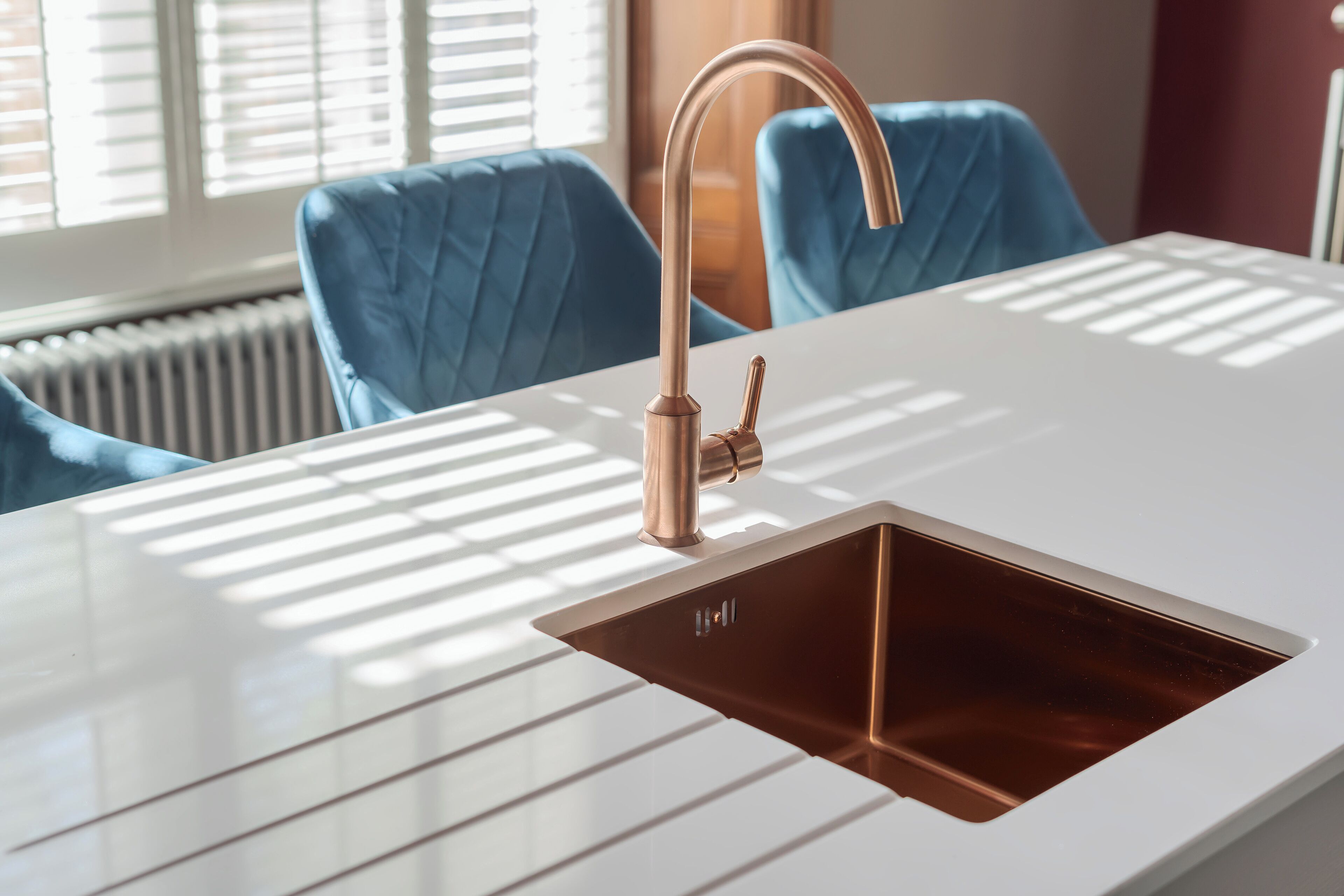 A modern kitchen with a copper faucet, white countertop, and blue upholstered chairs.