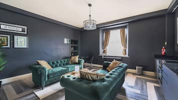 A modern living room with dark walls, a green sofa set, a chandelier, and a view of the outdoors through a window.
