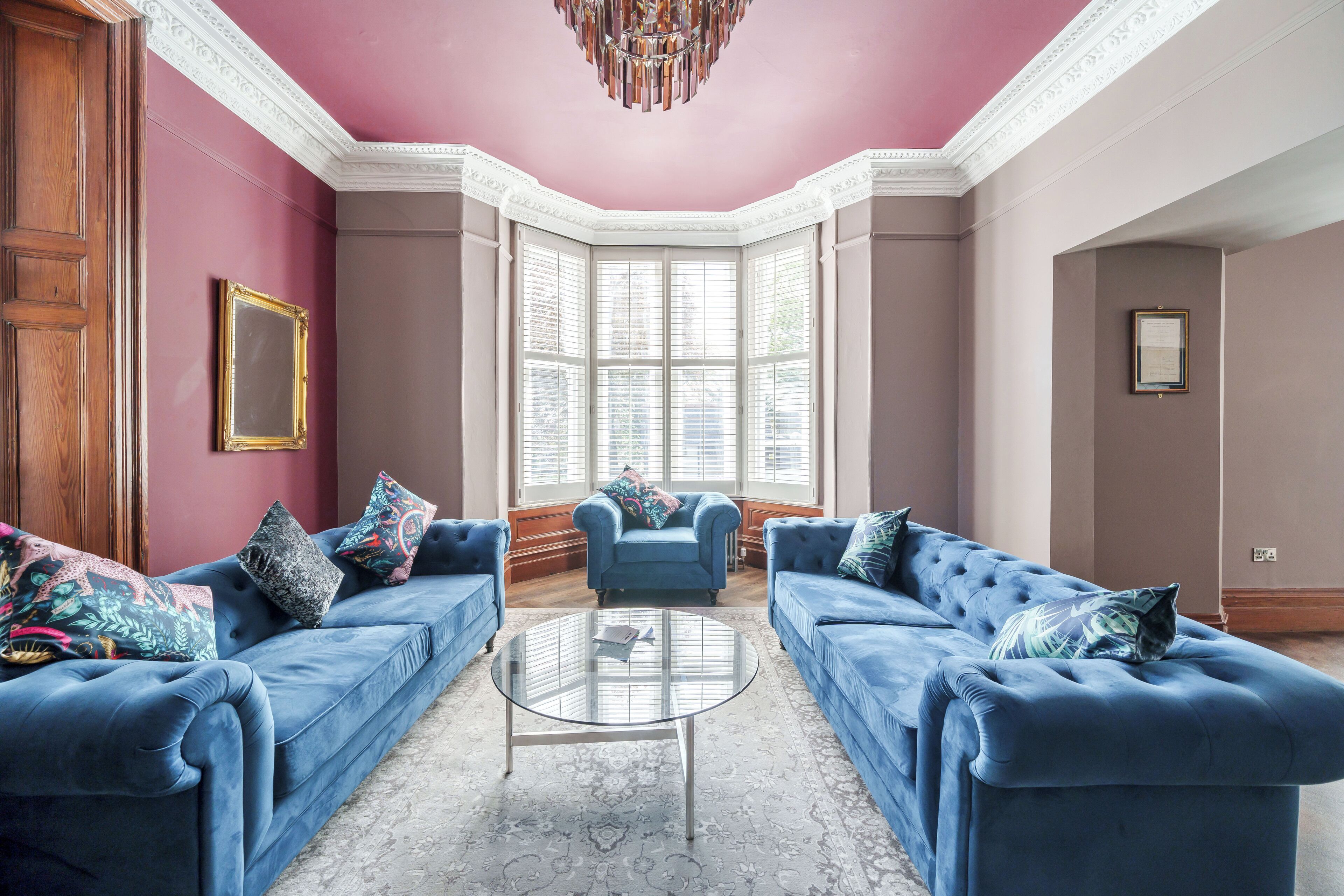Una habitación con dos sofás azules, una mesa de centro de vidrio y un ventanal con contraventanas blancas.