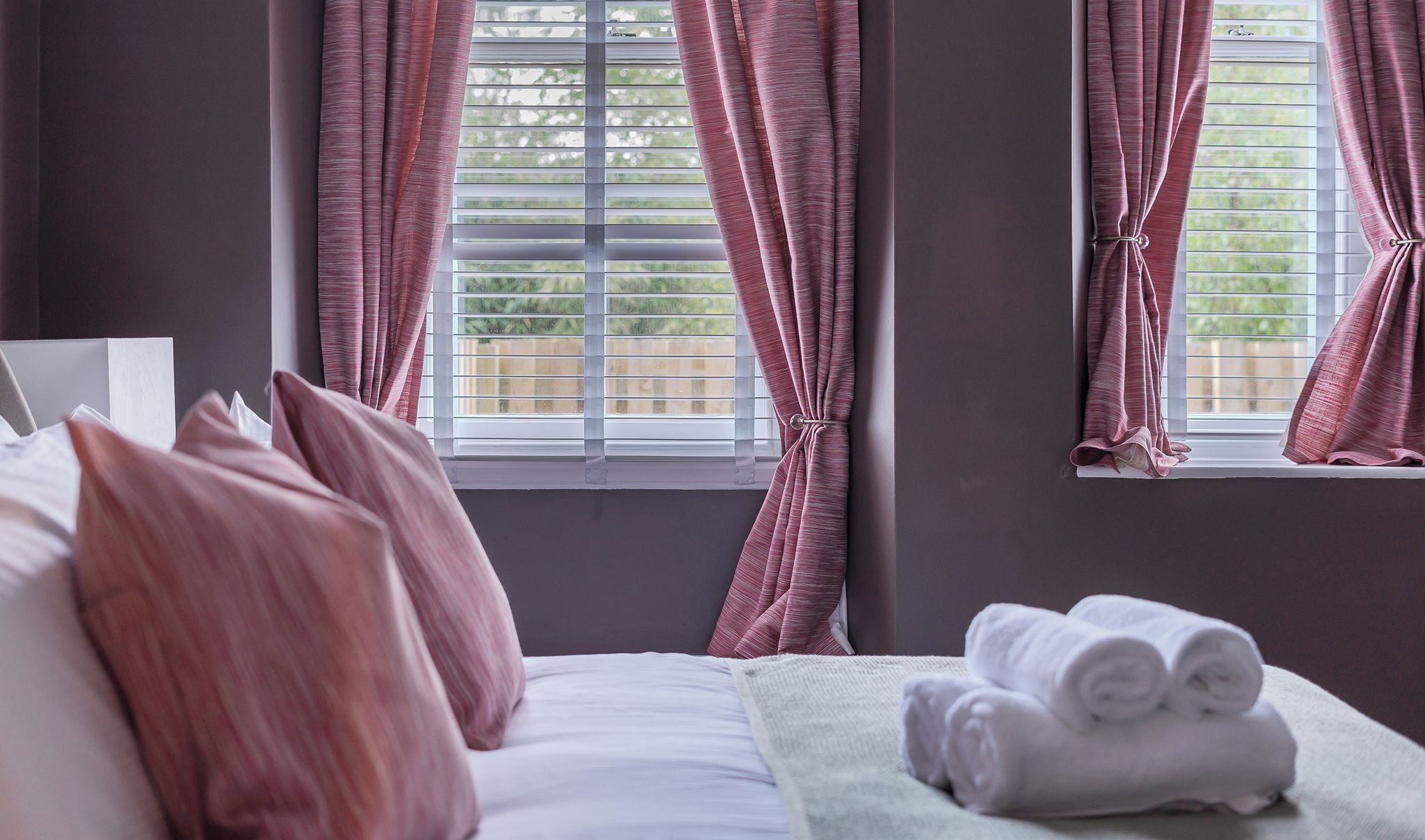 A Super King Size Bed with pink curtains, a window with blinds, and a stack of towels on the side.