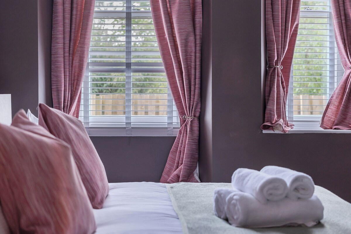 A Super King Size Bed with pink curtains, a window with blinds, and a stack of towels on the side.