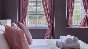 A Super King Size Bed with pink curtains, a window with blinds, and a stack of towels on the side.