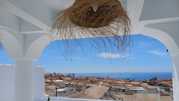 A balcony with a view of a coastal town, white columns, and a hanging basket. - House with 2 terraces and sea view. 2 Bedrooms, 500m from the Beach. (Puerto de Mazarrón)
