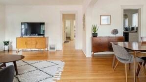 Una sala moderna con una mesa de comedor de madera, un sofá, un televisor sobre un mueble de madera y una cocina visible a través de un umbral abierto.