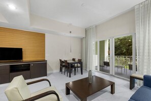 A modern living room with a flat-screen TV, a dining area with a table and chairs, and a balcony with a view of greenery. - Pool, Spa, Dining, Parking – Resort Style Studio (Miami)