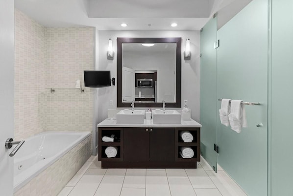 A modern bathroom with a bathtub, double sinks, a large mirror, and a flat-screen TV.