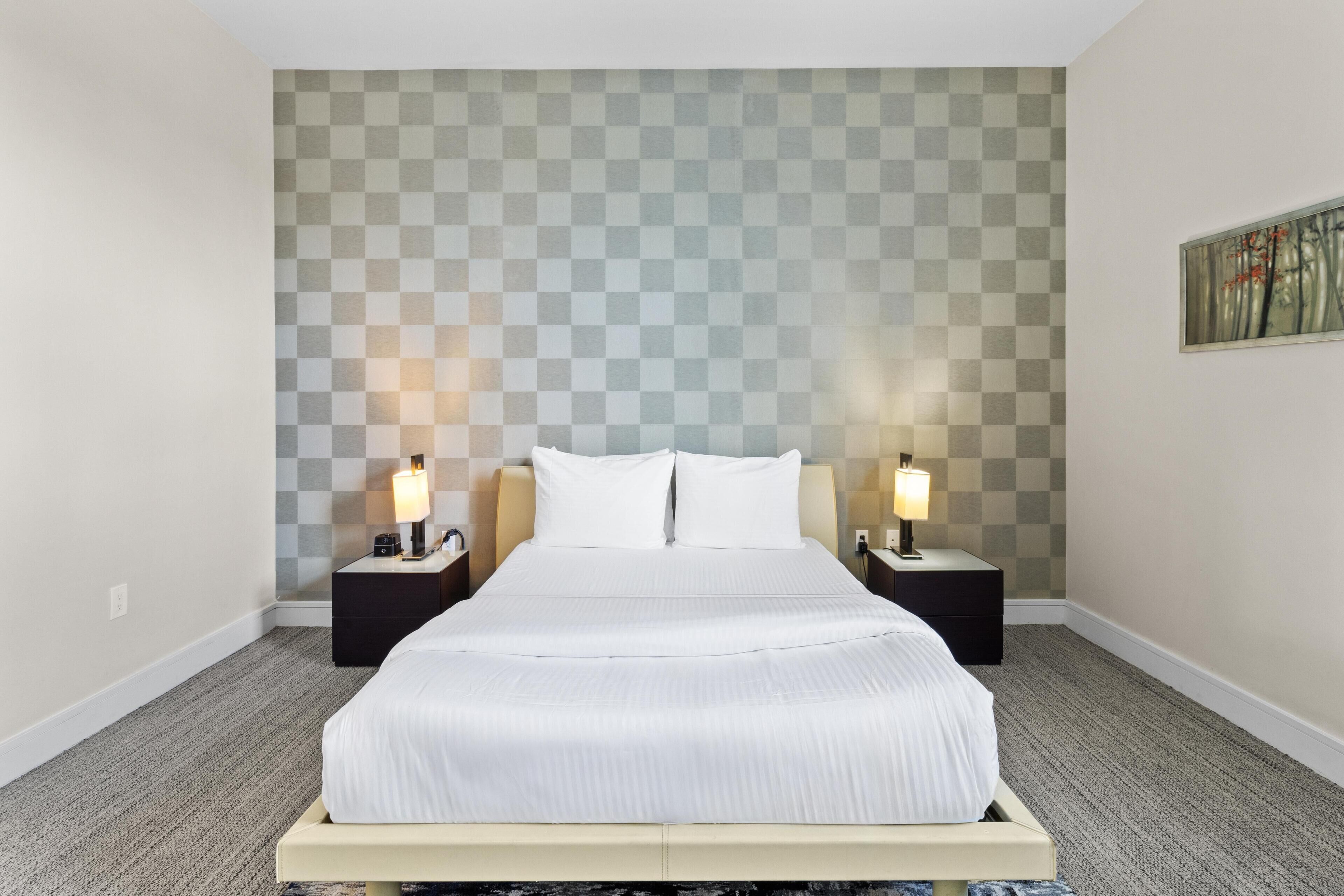 Une chambre d’hôtel avec un grand lit, deux tables de chevet avec des lampes, un motif à carreaux sur le mur et un tableau encadré accroché au mur.