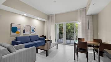A modern living room with a blue sofa, a patterned armchair, a dining table with chairs, and a balcony with a view of greenery.