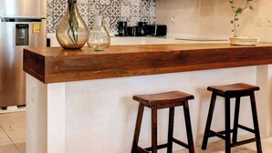 Une cuisine dotée d’un îlot central en bois, de deux tabourets de bar, d’un réfrigérateur, d’une machine à café et de luminaires suspendus.