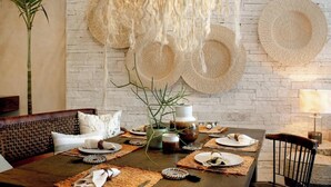Un coin repas avec une table en bois rustique mise pour le repas, entourée de sets de table en fibres tressées et d’une variété d’assiettes.