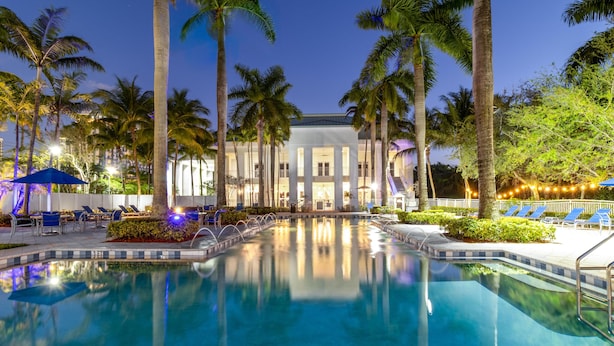 A pool area with lounge chairs, palm trees, and a building with columns. - Studio for 5, Downtown Doral, kitchen & parking (Miami)