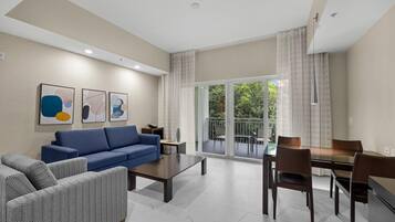A modern living room with a blue sofa, a patterned armchair, a dining table with chairs, and a balcony with a view of greenery.
