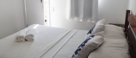 A neatly made bed with white linens and blue floral pillows, a wooden headboard, and a window with curtains.