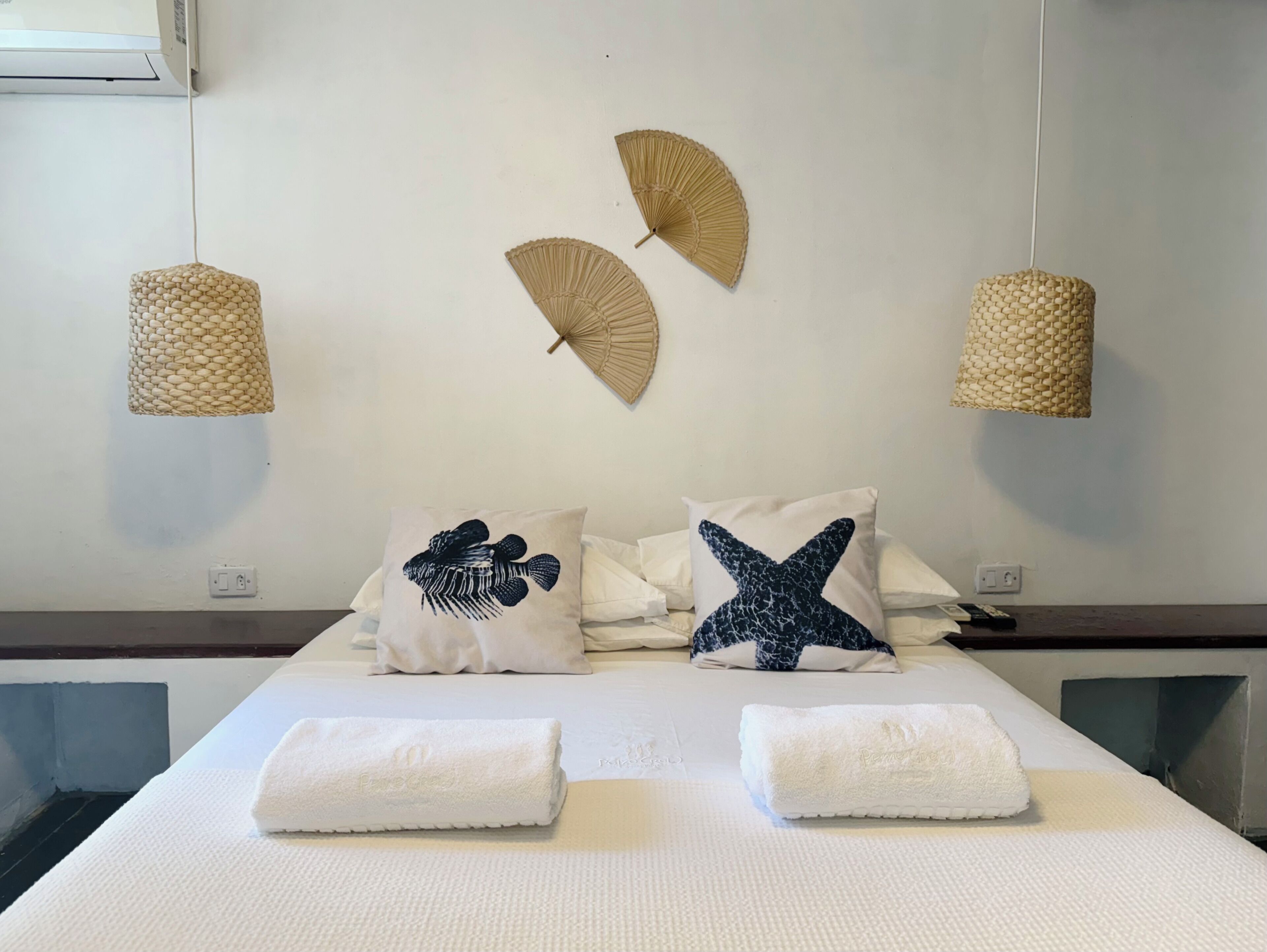 A hotel room with a bed, two pillows, two wall-mounted fans, and two towels.