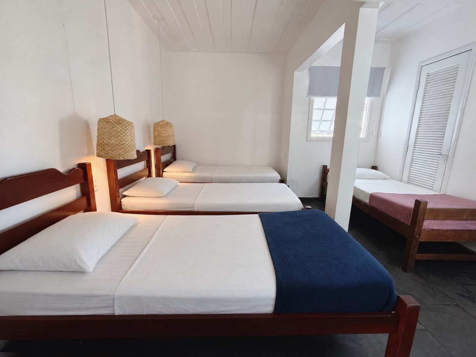 A room with four wooden beds arranged in a row, each with white bedding and a blue blanket on one bed. There are woven lamps on the headboards, and a window with blinds to the right.