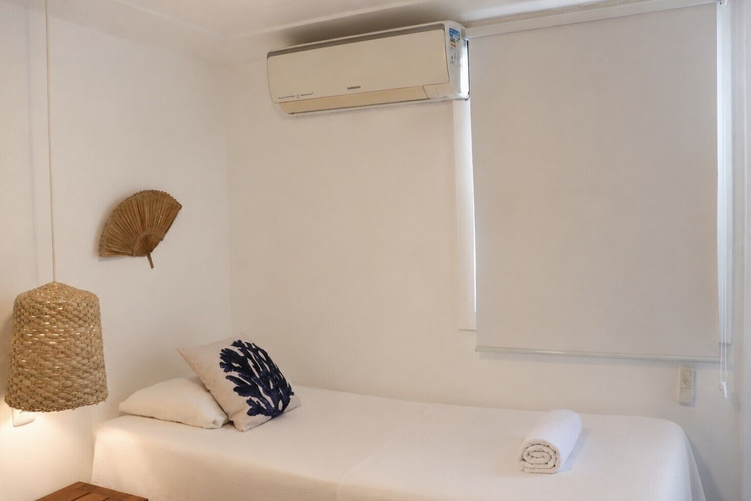 A bedroom with a bed, a wall-mounted air conditioner, a wall fan, and a bedside lamp.