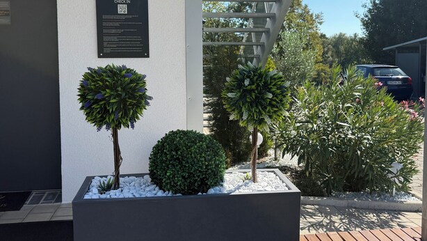 A building entrance with a QR code for check-in, a sign for Gastehaus Estenfeld, and two potted plants with white pebbles. - Gästehaus Würzburg 24h (Estenfeld)