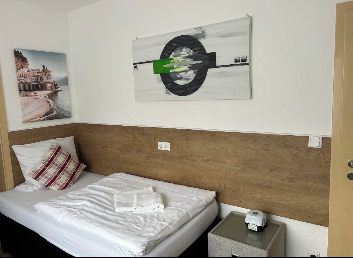 A hotel room with a bed, a wooden headboard, a wall-mounted painting, a nightstand, and a bedside table.