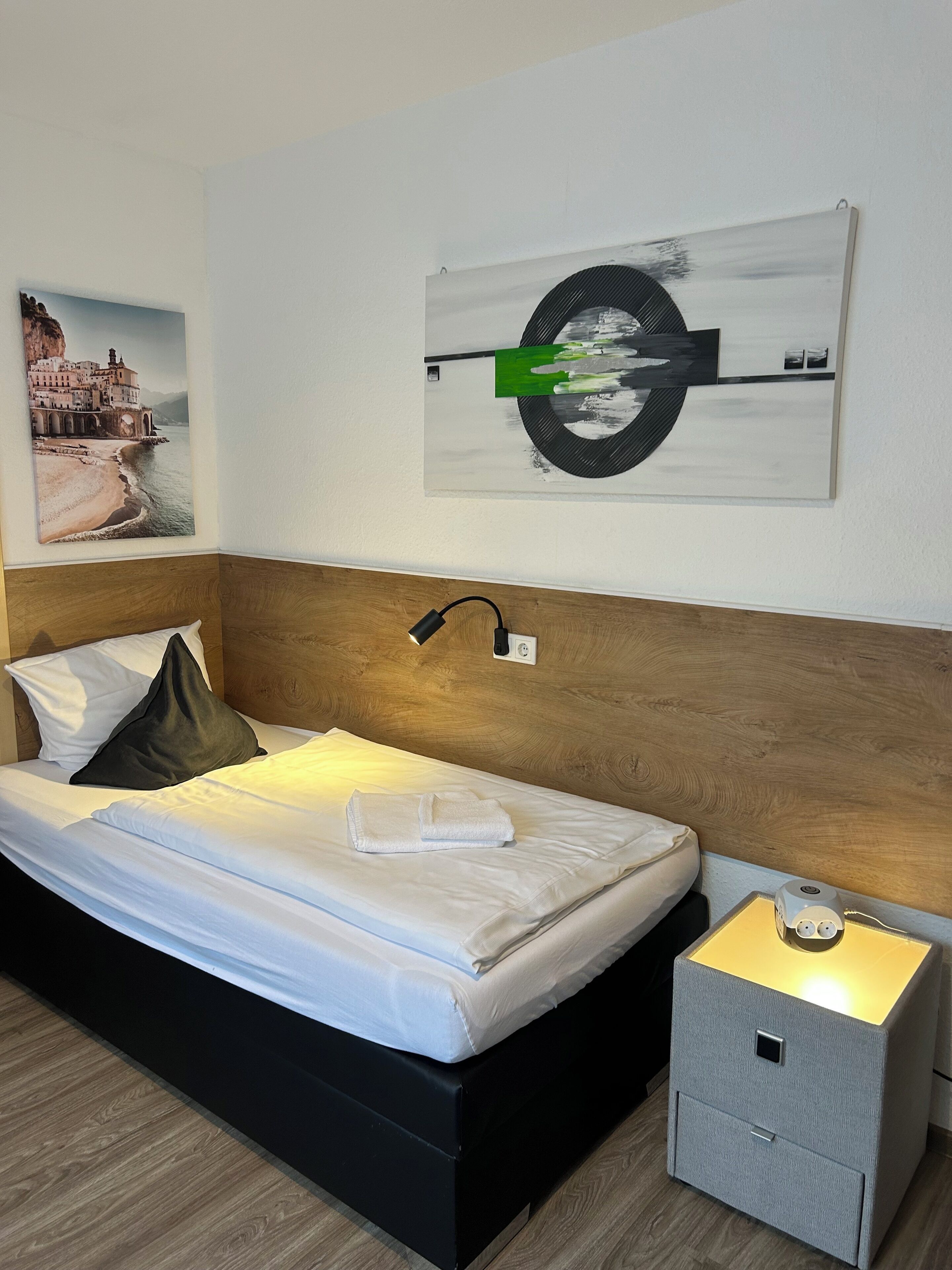 A hotel room with a bed, a nightstand, a lamp, a painting, and a small storage unit.