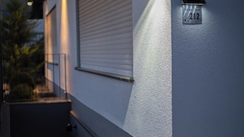 A modern building exterior with a number plaque, a wall-mounted light, and a window with blinds.