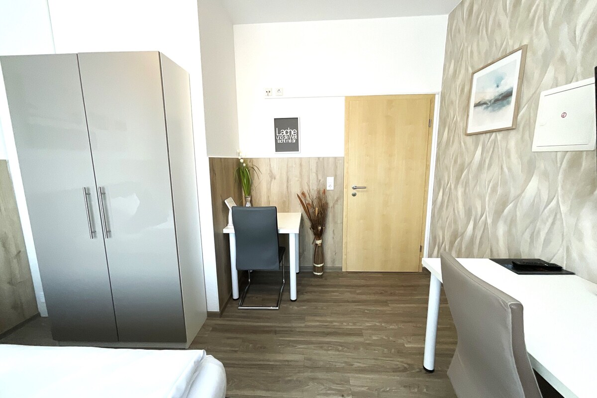 A modern hotel room with a bed, a desk, a chair, a wardrobe, and a door.