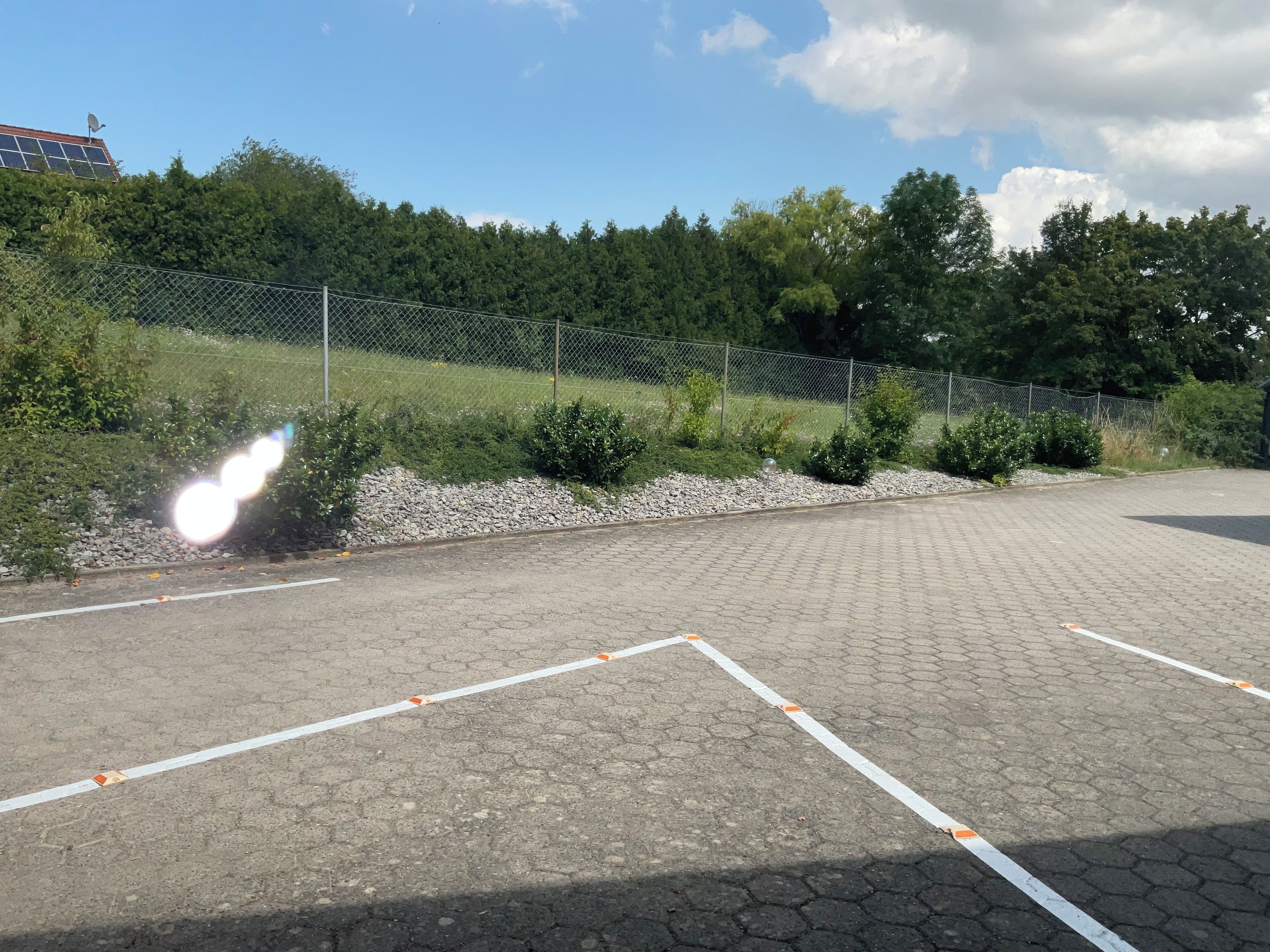 A paved parking area with white lines marking parking spaces, surrounded by a chain-link fence and greenery.