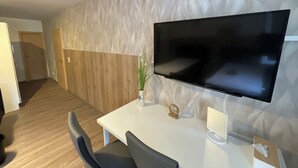 A modern hotel room with a large flat-screen TV mounted on the wall, a wooden accent wall, a white desk with a chair, and a small plant on the desk. - Gästehaus Würzburg 24h (Estenfeld)