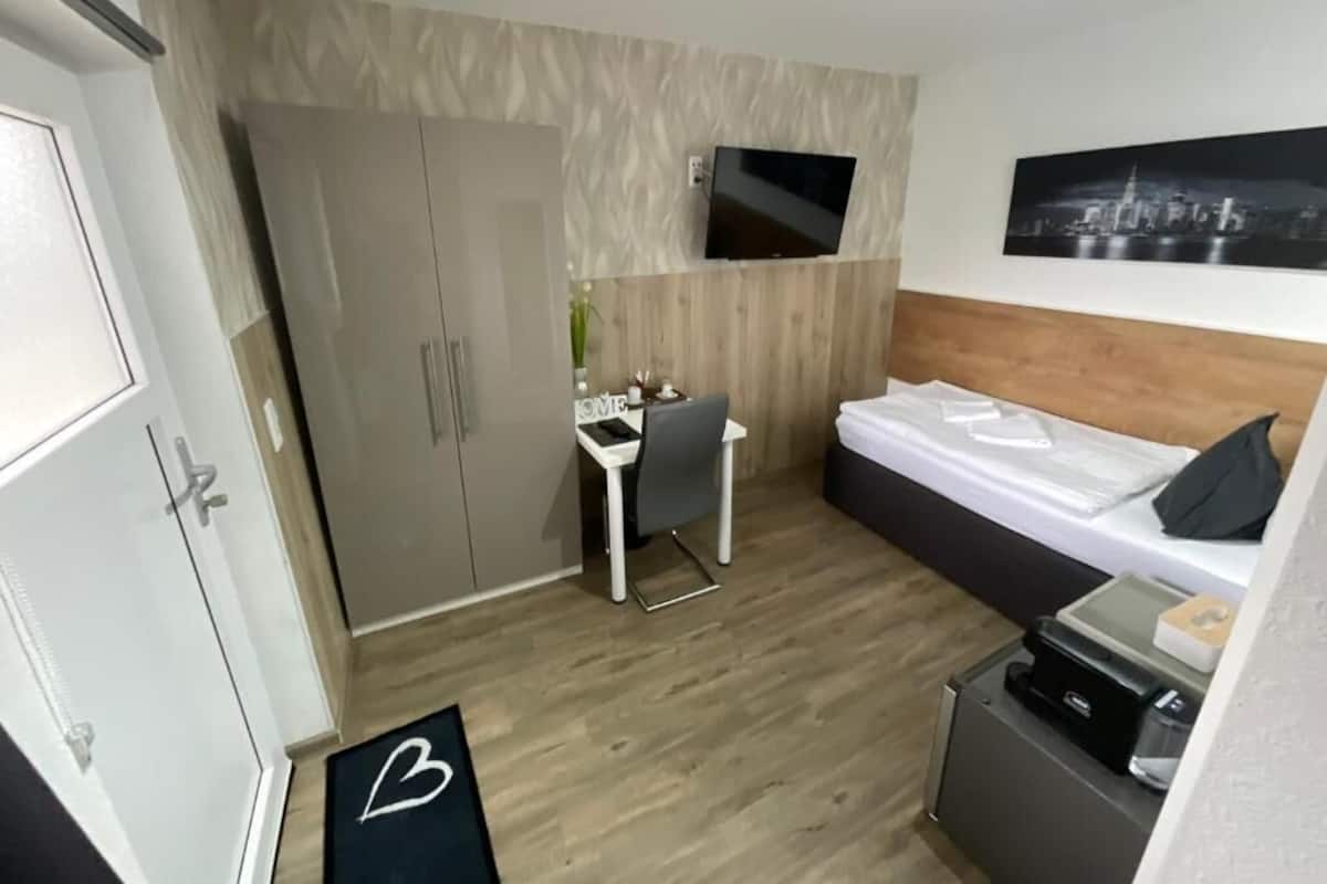 Une chambre d’hôtel de taille réduite, comprenant un lit, un bureau, une chaise, un téléviseur fixé au mur et une armoire.