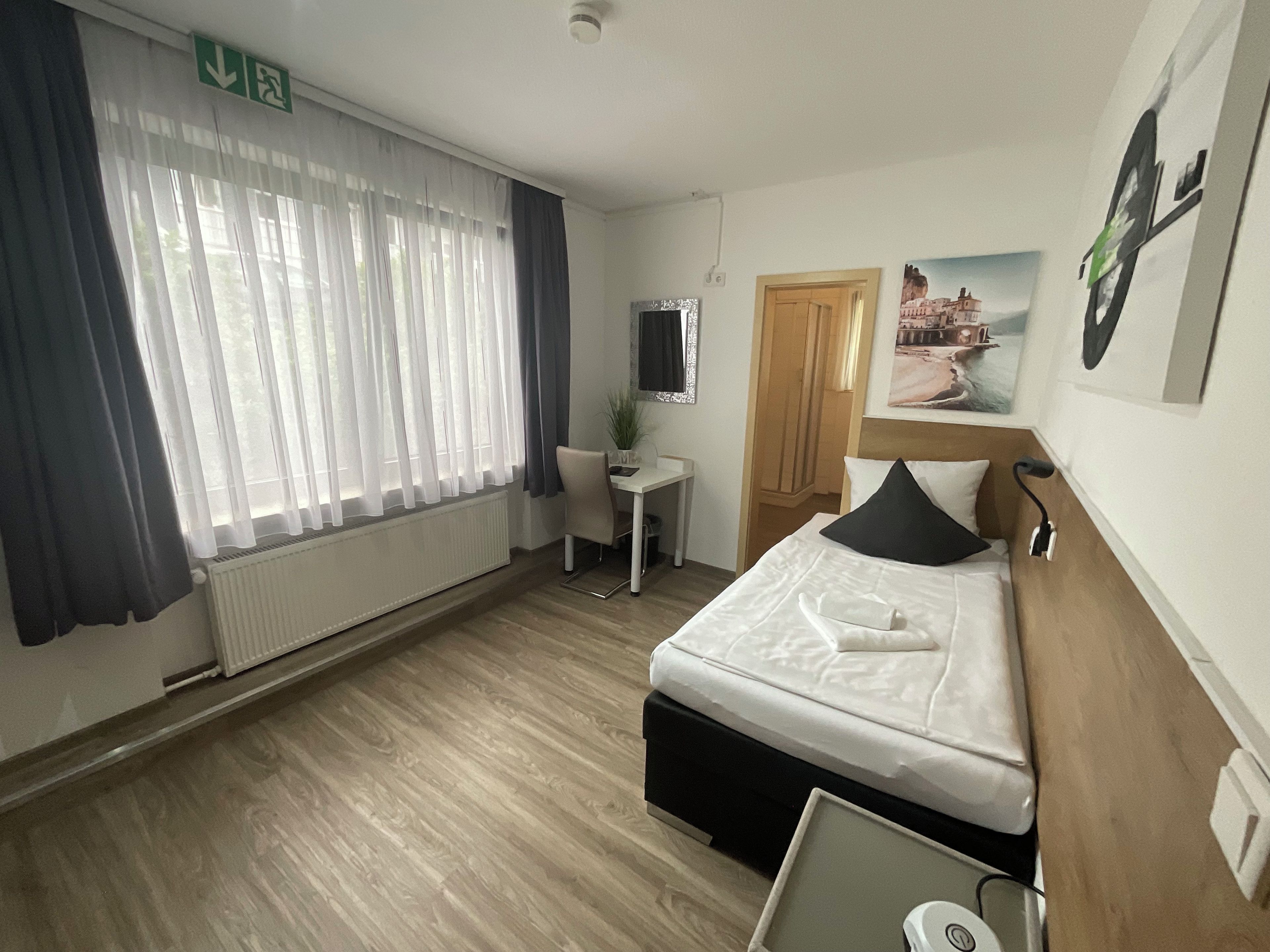 Ein Hotelzimmer mit einem Bett, einem Schreibtisch, einem Stuhl, einem Heizkörper, einem Fenster mit Vorhängen, einer Tür und einem an der Wand befestigten Fernseher.