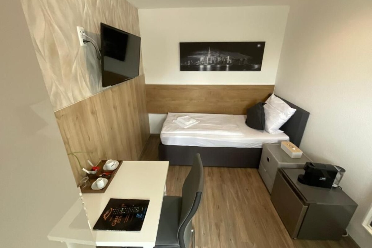 A compact hotel room with a bed, a desk with a chair, a small table with cups and a remote, a TV mounted on the wall, and a framed picture above the bed.