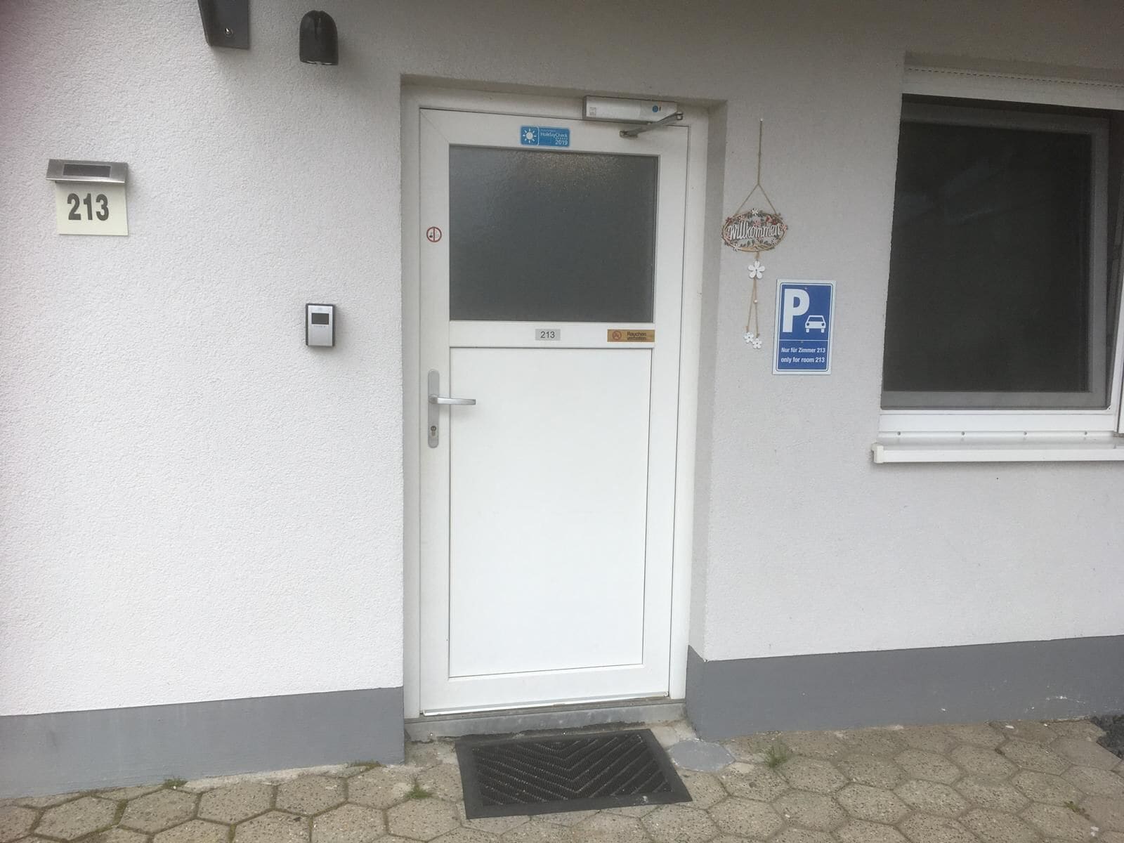 Puerta blanca con una placa que indica el número '213', un letrero de 'Bienvenido' y un letrero de 'Estacionamiento' que señala un lugar reservado para la habitación '213'.
