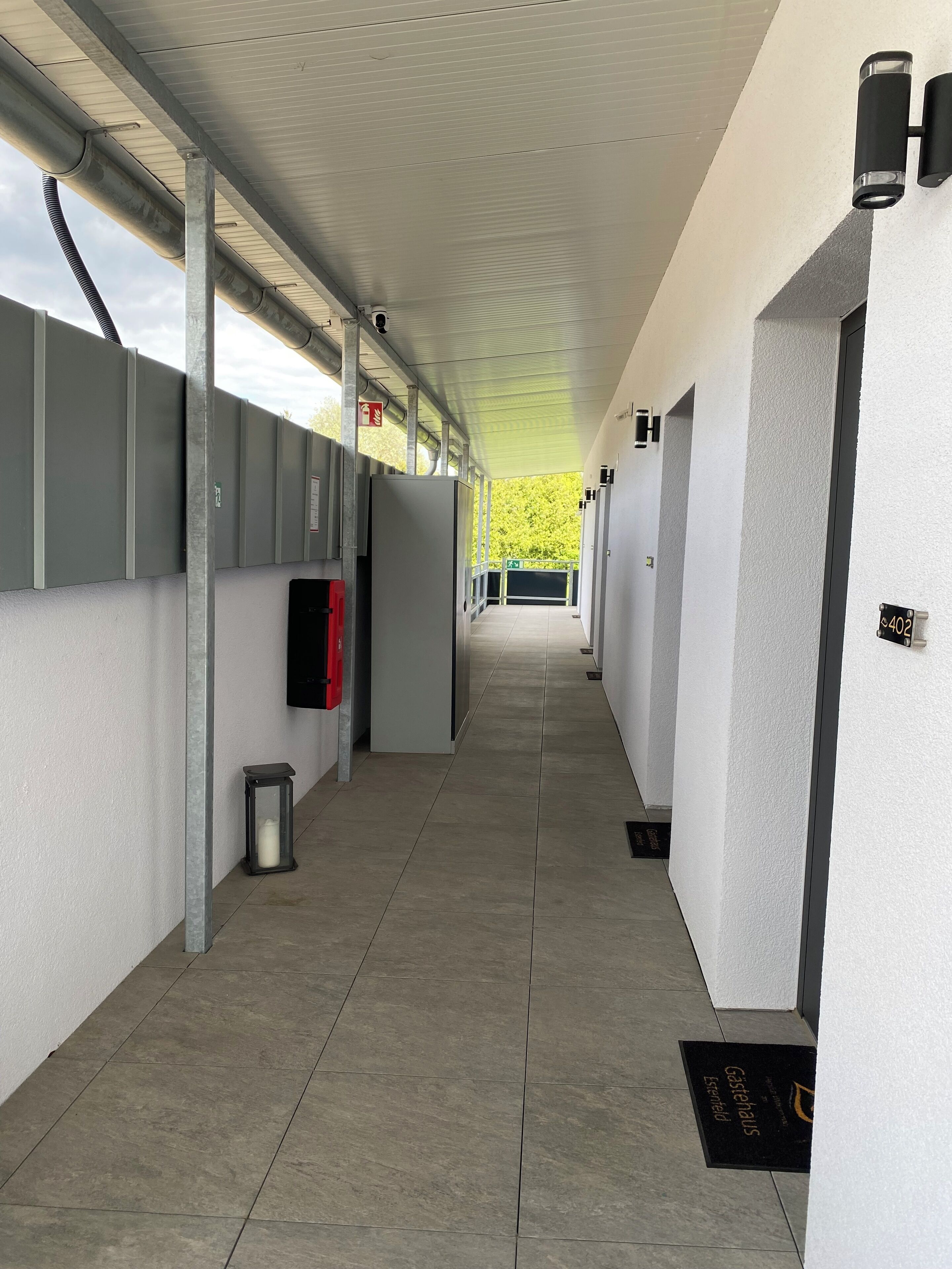 A corridor with a tiled floor, grey walls, and a ceiling with exposed pipes. There are doors on the right, a trash bin, and a black doormat with text.