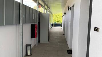 A corridor with a tiled floor, grey walls, and a ceiling with exposed pipes. There are doors on the right, a trash bin, and a black doormat with text.