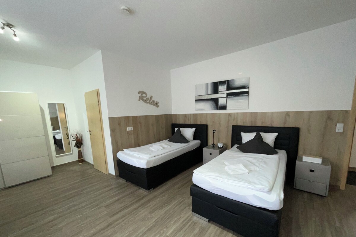 A modern hotel room with two beds, a wooden floor, a mirror, and a 'Relax' sign on the wall.