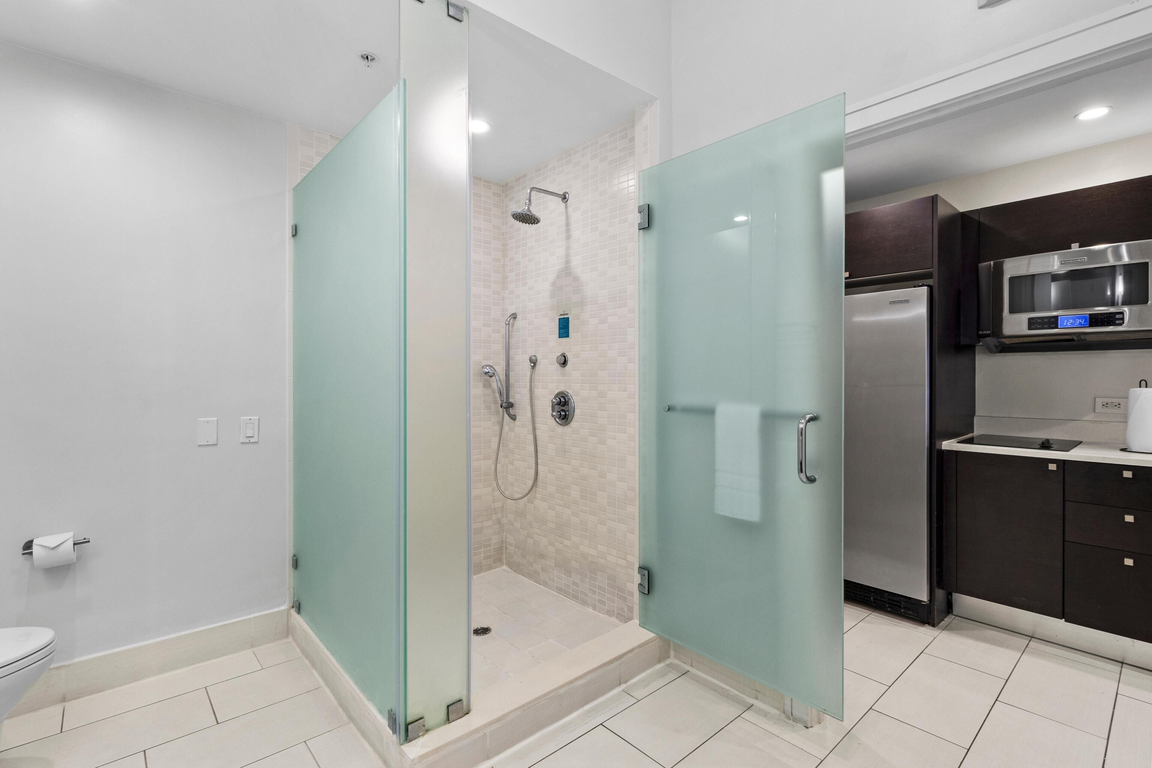 A modern bathroom with a glass-enclosed shower, a toilet, and a kitchen area with a refrigerator and microwave.