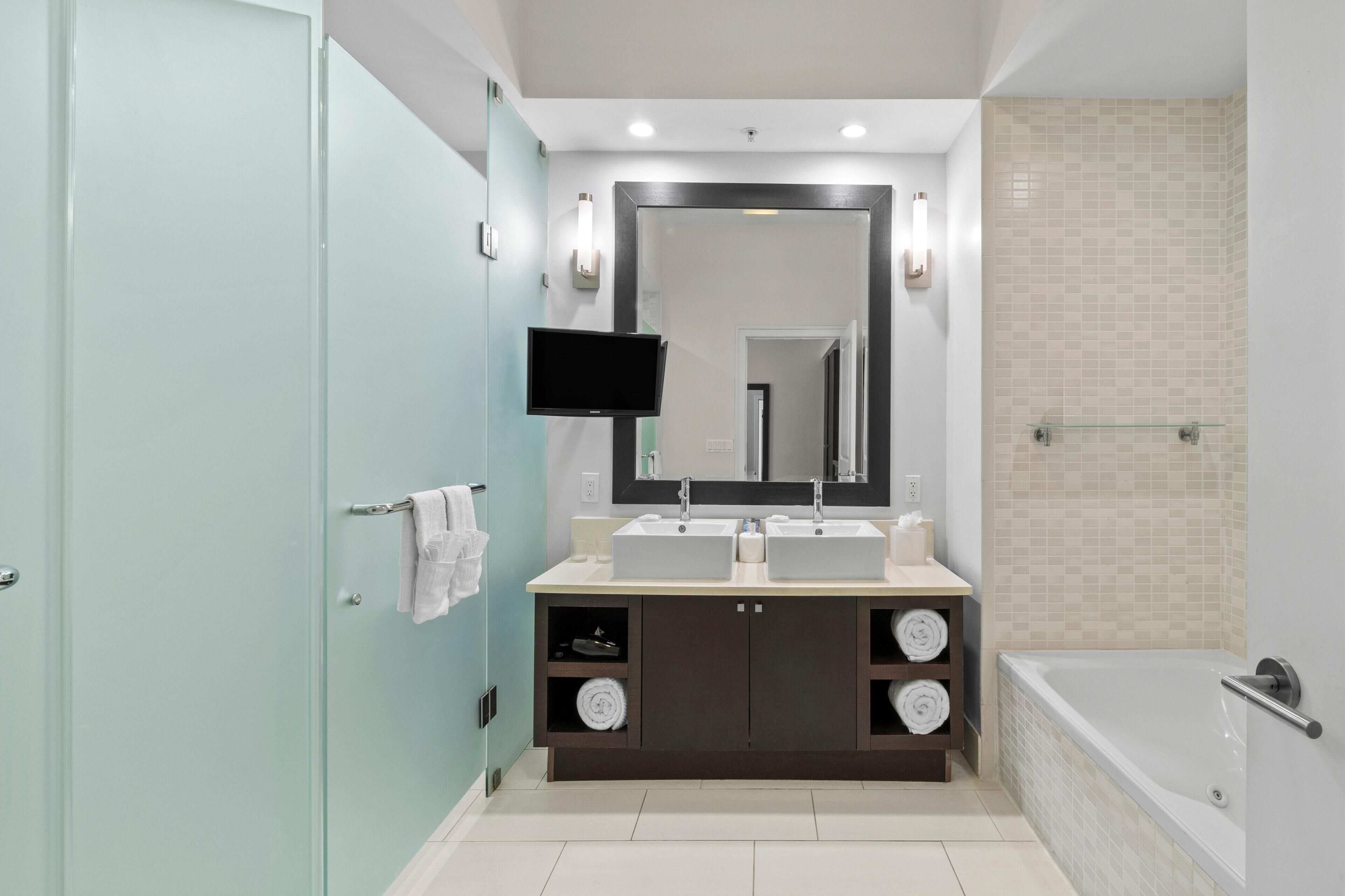 A modern bathroom with a dual sink vanity, a large mirror, a flat-screen TV, and a separate shower area.