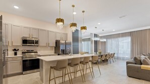 A modern kitchen with stainless steel appliances, a central island with bar stools, and a dining area with a grey sofa and curtains. - 12BR house with private pool in Paradiso Grande (Orlando)