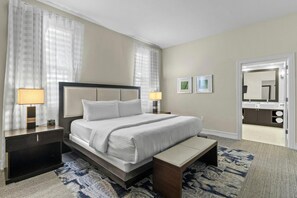 Habitación de hotel con una cama, mesitas de noche, un banco, un tapete y un baño visible a través de una puerta abierta.