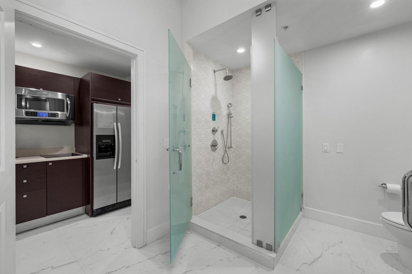 A modern bathroom with a glass-enclosed shower, a toilet, and a sink area with a stainless steel refrigerator.