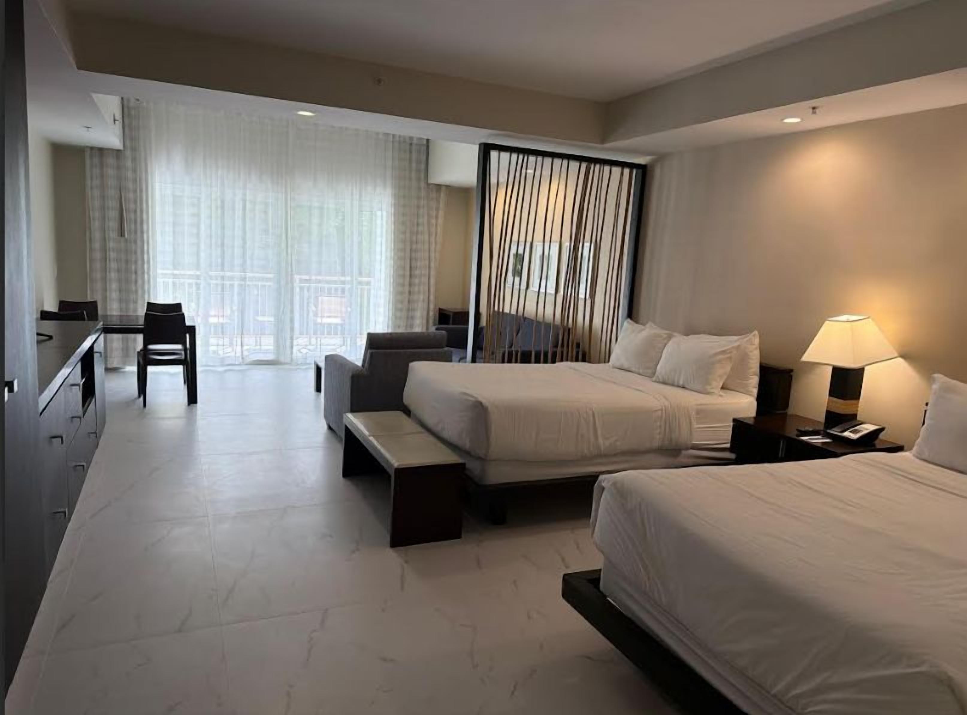 A hotel room with two beds, a desk, a chair, a sofa, and a large window with curtains.
