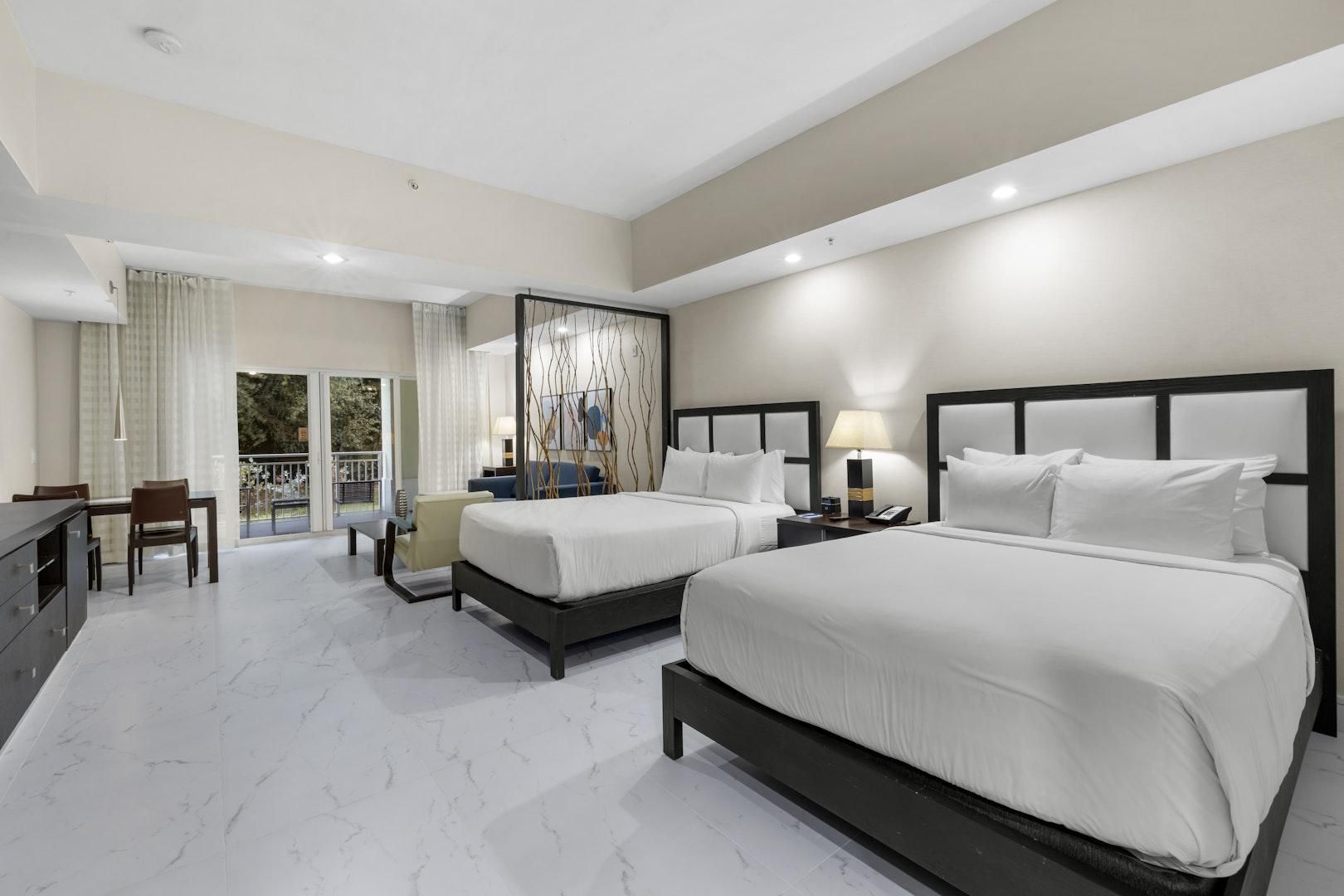 A hotel room with two beds, a dining area, and a balcony with curtains.