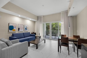 A modern living room with a blue sofa, a patterned armchair, a dining table with chairs, and a balcony with a view of greenery.