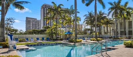 A swimming pool with lounge chairs and palm trees, surrounded by buildings.