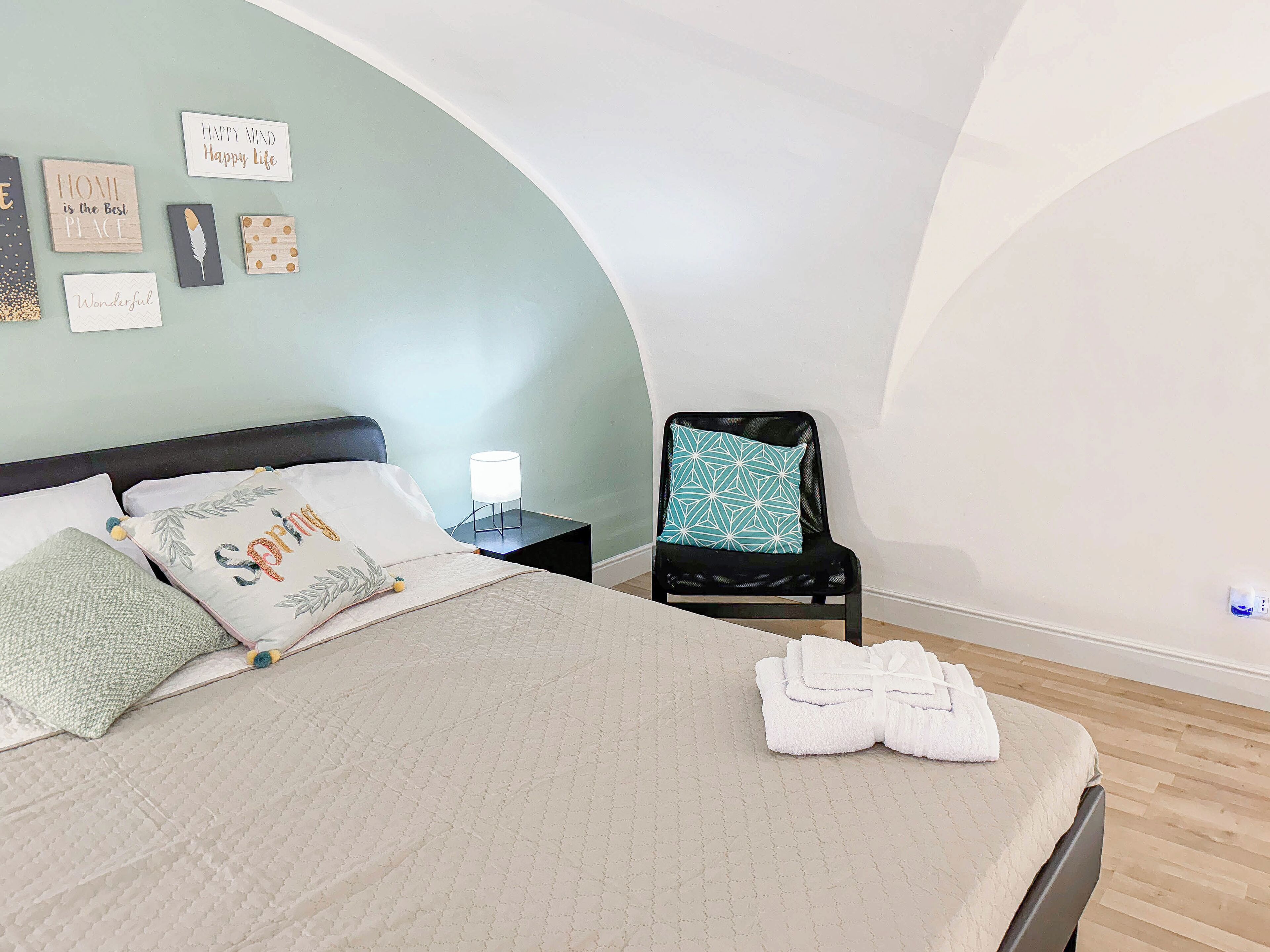 A neatly made bed with a white comforter and decorative pillows, a black chair with a patterned cushion, and a bedside table with a lamp.