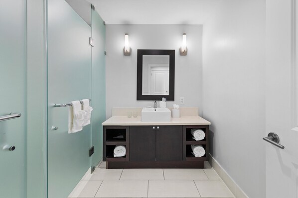 A modern bathroom with a glass shower enclosure, a dark vanity with a white sink, and two wall-mounted lights.