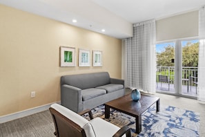 A living room with a gray sofa, a wooden coffee table, and a patterned rug. There are three framed pictures on the wall and a window with a view of greenery. - Relax & Unwind in Doral, Large Studio – sleep 5 (Miami)